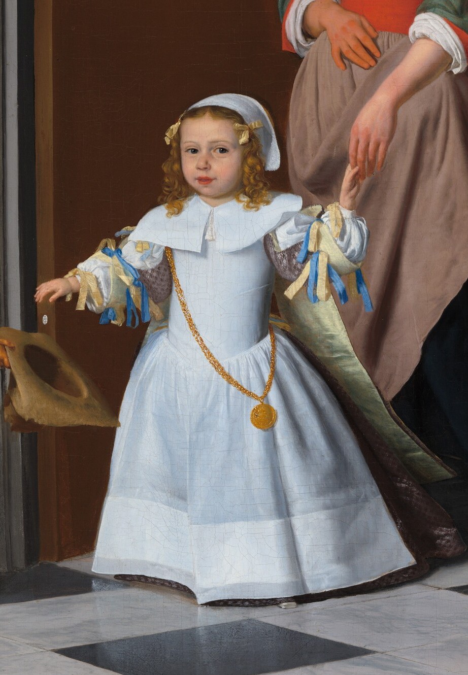 The child, in an elaborate white dress, reaches for a hat.