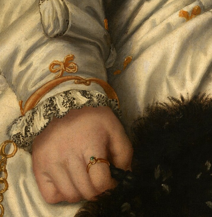 Her hands, adorned with rings, rest on rich textures.